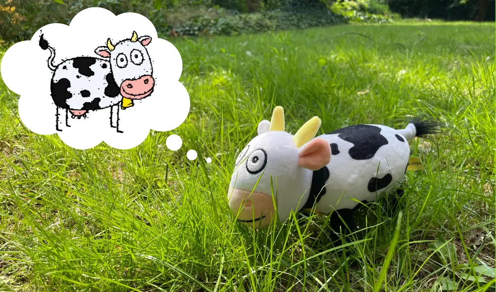 A stuffed cow toy lies on green grass, imagining a cartoon cow in a thought bubble above its head.